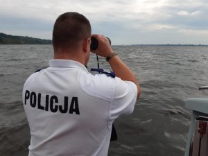 Zdjęcie pokazuje policjanta ogniwa wodnego obserwującego przez lornetkę regaty na wodzie