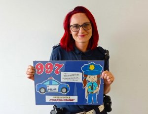 Policjantka stojąca na tle ściany i trzymająca laurkę otrzymaną od przedszkolaków. Laurka przedstawia napis 997, radiowóz i podziękowanie