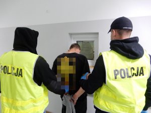 Zdjęcie przedstawia zatrzymanego mężczyznę do czynnej napaści na funkcjonariuszy prowadzonego korytarzem komendy przez dwóch policjantów