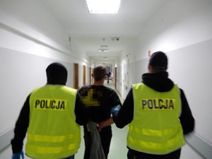 Zdjęcie przedstawia zatrzymanego mężczyznę do czynnej napaści na funkcjonariuszy prowadzonego korytarzem komendy przez dwóch policjantów