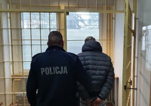 Po środku zdjęcia policjant z lewej strony natomiast po prawej zatrzymany kierowca. W tle widoczne kraty i okno. Zdjęcie do treści informacji