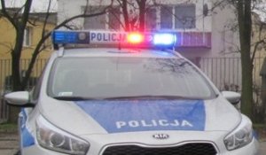 Pokazany od przodu policyjny radiowóz z maską koloru srebnego po bokach, a na środku na niebieskim tle napis POLICJA. U góry na dachu policyjny kogut z włączonymi światłami błyskowymi