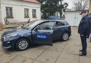 Radiowóz kolory ciemny niebieski stojący na tle budynku. Ma otwarte drzwi na których jest napis policja. Na dachu dwa koguty policyjne koloru niebieskiego i czerwonego. Od prawej strony stoi policjant.