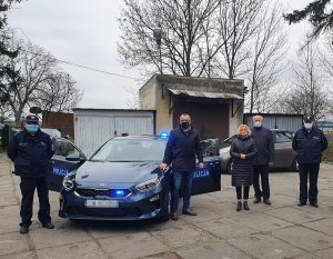 Po lewej stronie zdjęcia stoi policjant, następnie przekazywany radiowóz, Po prawej stronie stoją cztery osoby zgodnie z treścią informacji