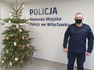 Dzielnicowy stojący po prawej stronie zdjęcia na tle ściany z napisem Komenda Miejska Policji we Włocławku oraz po lewej stronie choinka