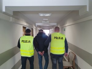 Policjanci podczas doprowadzania osoby poszukiwanej. Z lewej i prawej strony policjanci w żółtych kamizelkach, a po środku zatrzymany mężczyzna zgodnie z treścią komunikatu