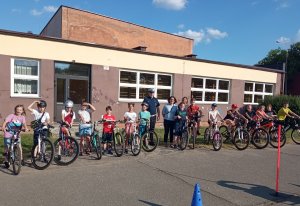 Grupa uczniów uczestniczących w egzaminie na kartę rowerową. Po środku umundurowany policjant. Wszyscy stoją na tle budynku szkoły