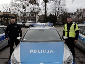 Policjanci umundurowani stoją po lewej i prawej stronie radiowozu. Radiowóz widoczna maska przednia z napisem policja i policyjne sygnały uprzywilejowania