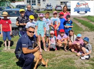 Dzieci podczas wizyty w komendzie. Dzieci stoją przodem, a przed nimi siedzi policjant z psem słuzbowym