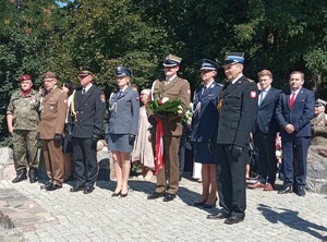 przedstawiciele różnych służb mundurowych stoją obok siebie, mężczyzna w mundurze wojskowym trzyma wiązankę kwiatów