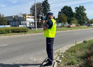 policjant stoi przy drodze i trzymanym w ręku urządzeniem mierzy prędkość pojazdów