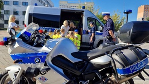 Policjanci prezentują dzieciom ubranym w kamizelki odblaskowe radiowóz, motocykl, quad i inny sprzęt wykorzystywany podczas służby