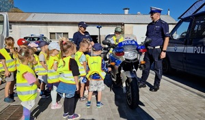 Policjanci prezentują dzieciom ubranym w kamizelki odblaskowe radiowóz, motocykl, quad i inny sprzęt wykorzystywany podczas służby