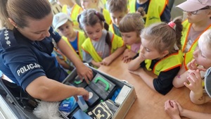 Dzieci ubrane w kamizelki odblaskowe oglądają policyjny radiowóz, motocykl, guad i walizkę technika kryminalistyki
