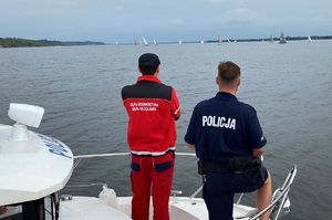 widać pływające w oddali żaglówki, a na pierwszym planie widoczny fragment łodzi z napisem policja oraz stojący na niej policjant i ratownik WOPR