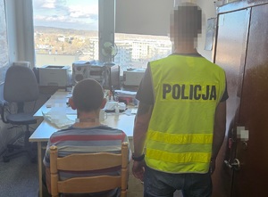 Policjanci podczas czynności z zatrzymanymi osobami w pomieszczeniu jednostki. Po prawej stoi policjant w żółtej kamizelce z napisem policja, po lewej siedzą tyłem osoby