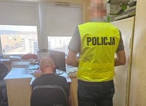 Policjanci podczas czynności z zatrzymanymi osobami w pomieszczeniu jednostki. Po prawej stoi policjant w żółtej kamizelce z napisem policja, po lewej siedzą tyłem osoby