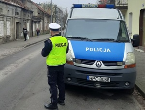 Policjant stoi przy radiowozie i patrzy na zachowania kierujących i pieszych w rejonie drogi