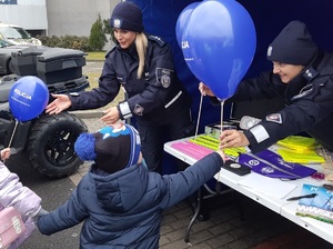 Dzieci i dorośli stoją przy policyjnym stoisku. Obok stoika stoi policyjny radiowóz i quad. Na stoisku leżą balony, odblaski, gogle, gazetki i ulotki policyjne.