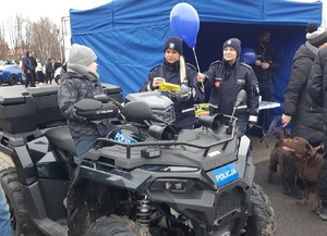 Dzieci i dorośli stoją przy policyjnym stoisku. Obok stoika stoi policyjny radiowóz i quad. Na stoisku leżą balony, odblaski, gogle, gazetki i ulotki policyjne.