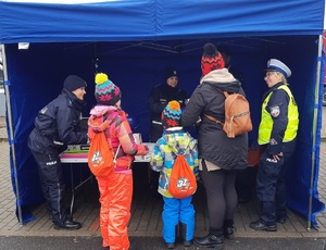 Dzieci i dorośli stoją przy policyjnym stoisku. Obok stoika stoi policyjny radiowóz i quad. Na stoisku leżą balony, odblaski, gogle, gazetki i ulotki policyjne.