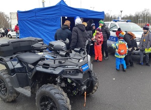 Dzieci i dorośli stoją przy policyjnym stoisku. Obok stoika stoi policyjny radiowóz i quad. Na stoisku leżą balony, odblaski, gogle, gazetki i ulotki policyjne.