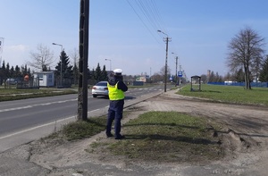 Policjant w żółtej kamizelce odblaskowej z napisem POLICJA stoi przy drodze i mierzy prędkość
