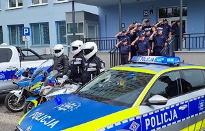 Policjanci oddają hołd przed komendą, stoją radiowozy z włączonymi sygnałami uprzywilejowania