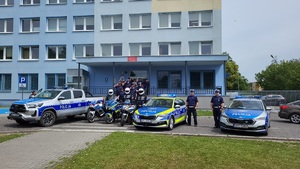 Policjanci oddają hołd przed komendą, stoją radiowozy z włączonymi sygnałami uprzywilejowania