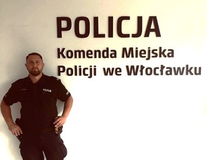 Policjant stojący na tle ściany z napisem Komenda Miejska Policji we Włocławku