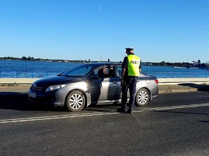 Policjant w kamizelce odblaskowej z napisem POLICJA stoi na jezdni przy zatrzymanym aucie i kontroluje kierowcę