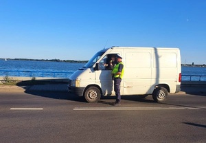 Policjant w kamizelce odblaskowej z napisem POLICJA stoi na jezdni przy zatrzymanym aucie i kontroluje kierowcę