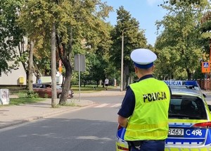 Policjant stojący tyłem  obserwuje skrzyżowanie. Przed nim stoi policyjny radiowóz