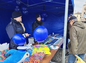 Policjanci i mieszkańcy przy stoisku promocyjnym. Na stołach leżą policyjne smycze, balony, odblaski, kolorowanki, gazety i inne gadżety. Przy stołach stoją policyjne pojazdy - dwa radiowozy i quad, na który wsiadają dzieci.