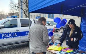 Policjanci i mieszkańcy przy stoisku promocyjnym. Na stołach leżą policyjne smycze, balony, odblaski, kolorowanki, gazety i inne gadżety. Przy stołach stoją policyjne pojazdy - dwa radiowozy i quad, na który wsiadają dzieci.