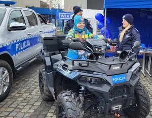 Policjanci i mieszkańcy przy stoisku promocyjnym. Na stołach leżą policyjne smycze, balony, odblaski, kolorowanki, gazety i inne gadżety. Przy stołach stoją policyjne pojazdy - dwa radiowozy i quad, na który wsiadają dzieci.