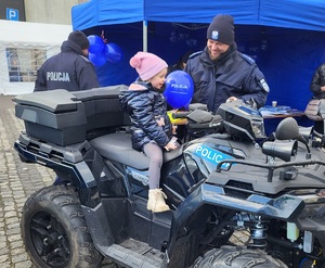 Policjanci i mieszkańcy przy stoisku promocyjnym. Na stołach leżą policyjne smycze, balony, odblaski, kolorowanki, gazety i inne gadżety. Przy stołach stoją policyjne pojazdy - dwa radiowozy i quad, na który wsiadają dzieci.