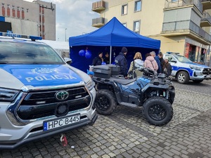 Policjanci i mieszkańcy przy stoisku promocyjnym. Na stołach leżą policyjne smycze, balony, odblaski, kolorowanki, gazety i inne gadżety. Przy stołach stoją policyjne pojazdy - dwa radiowozy i quad, na który wsiadają dzieci.