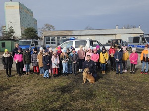 Policjanci podczas wizyty dzieci w komendzie. Policjant wraz z psem służbowym pokazuje tresurę i omawia z dziećmi zasady bezpieczeństwa